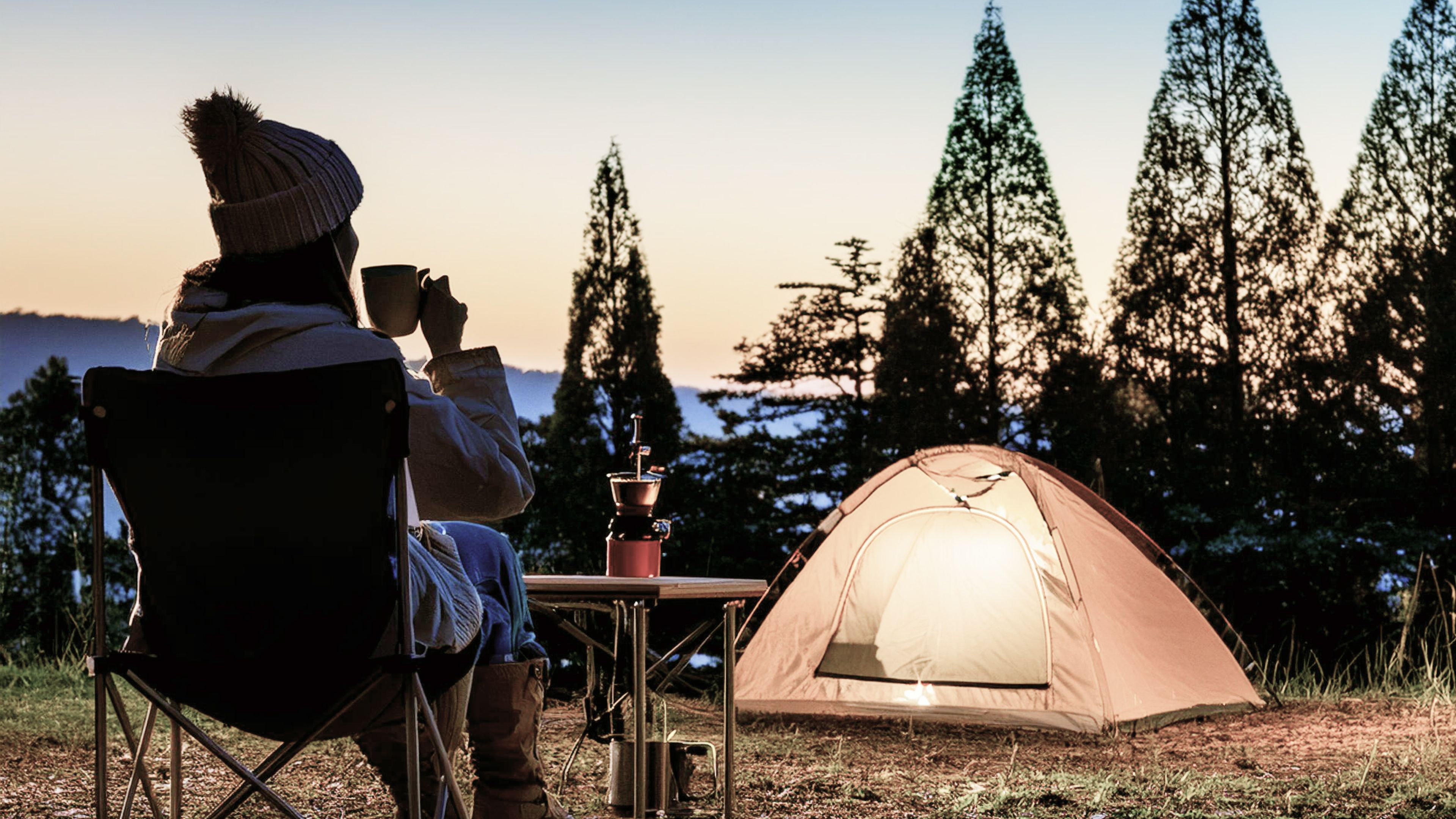 夕暮れ時、ベージュのテントの前で折りたたみチェアに座り、ソロキャンプを楽しみながらコーヒーを飲む女性